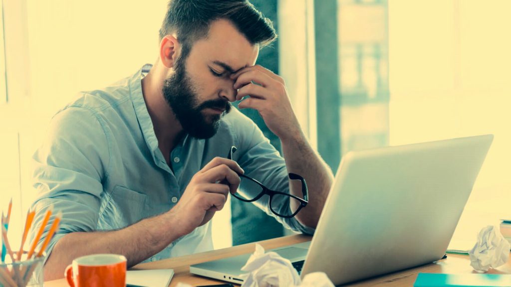 A frustrated young man with a beard, wearing a light blue shirt, sits at a desk with a laptop in front of him.