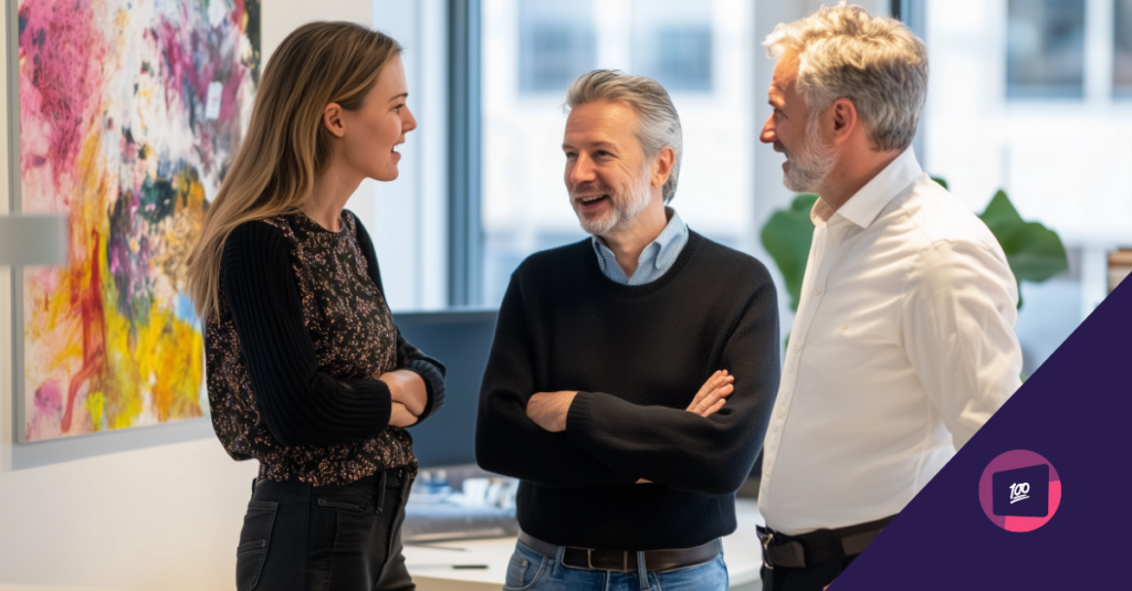 Three professionals engaged in a casual conversation in an office setting, with a colorful abstract painting in the background